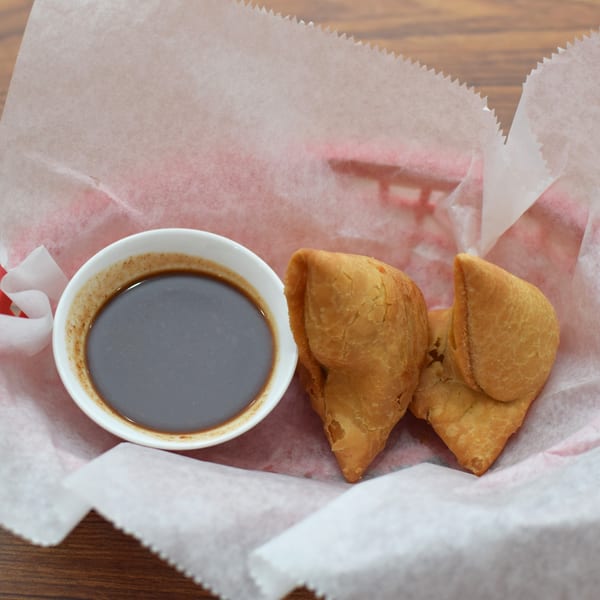 Two golden-brown samosas and a small white bowl of dark dipping sauce in a paper-lined basket.
