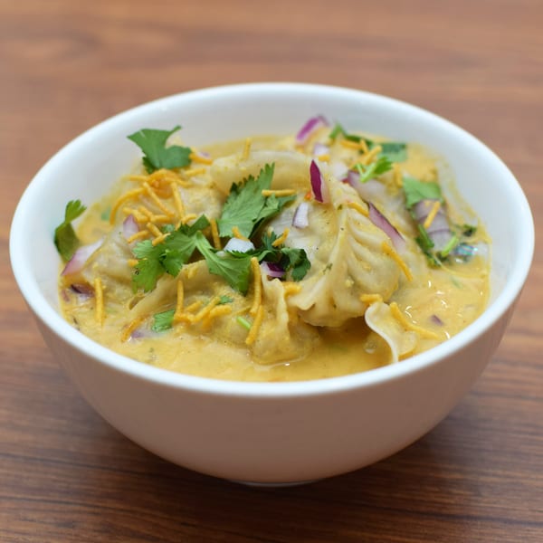 Indian momos in a creamy yellow sauce, garnished with red onions, cilantro, and crispy noodles.