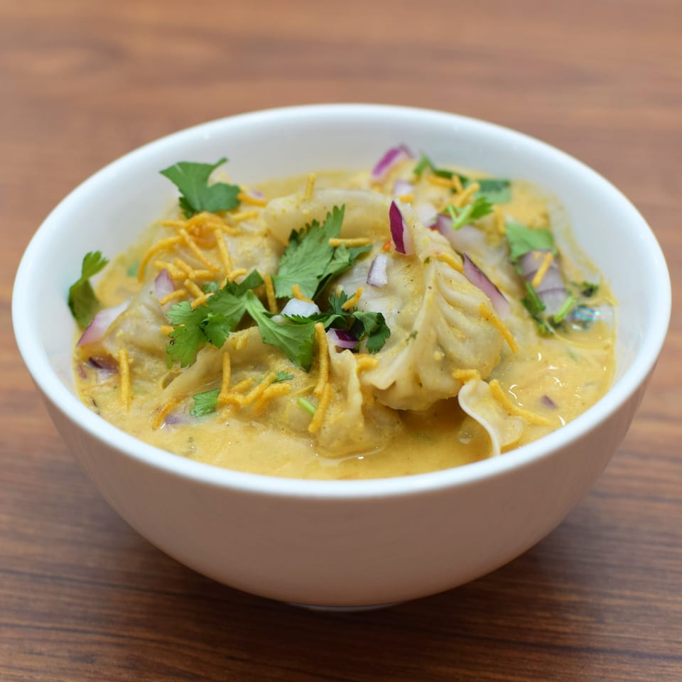 Indian momos in a creamy yellow sauce, garnished with red onions, cilantro, and crispy noodles.