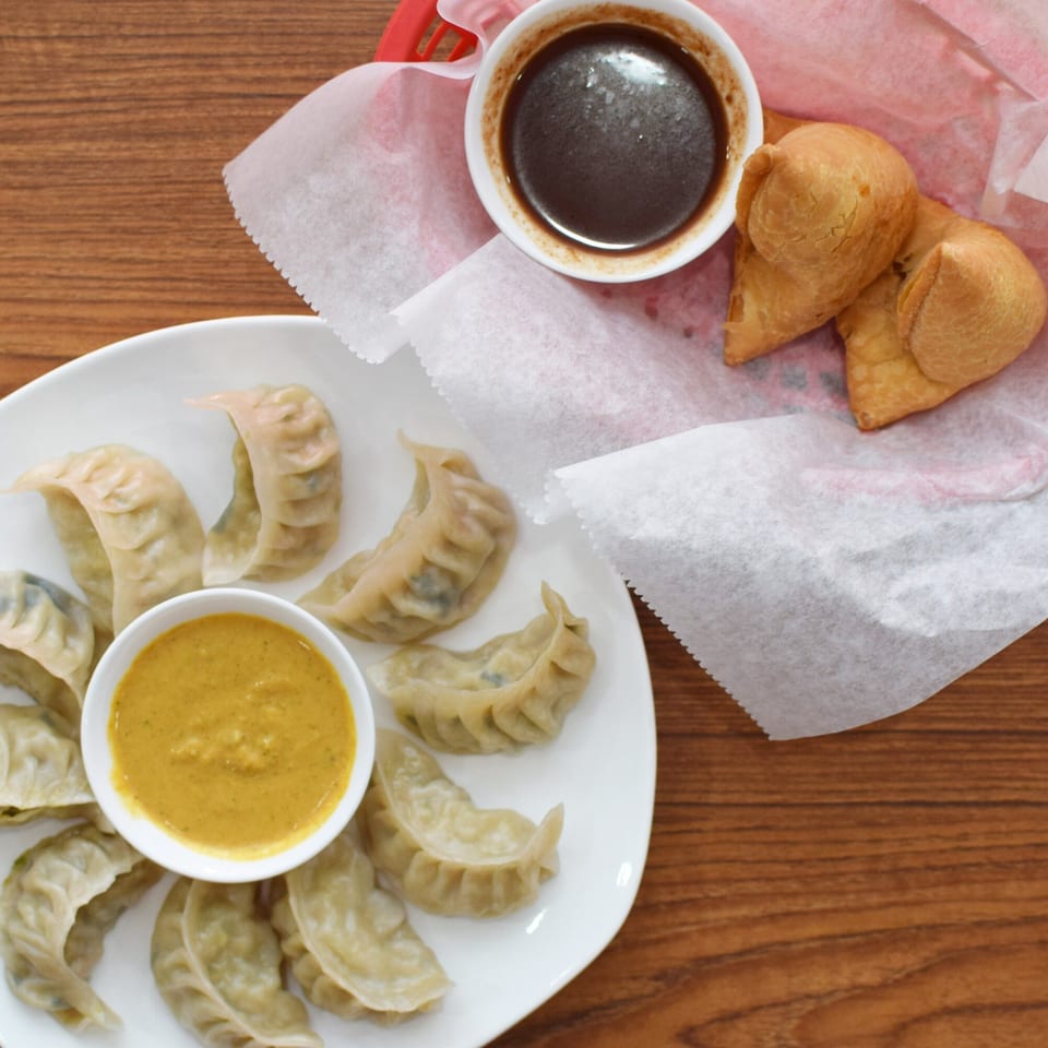 Overhead view of steamed momos with yellow dipping sauce, and samosas with dark sauce.