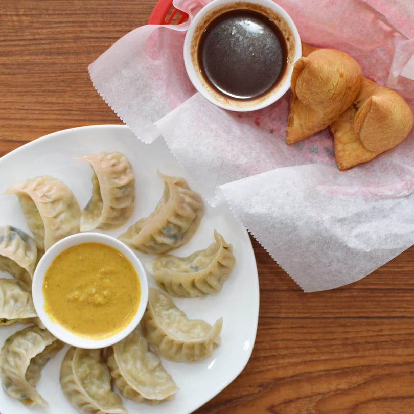 Overhead view of steamed momos with yellow dipping sauce, and samosas with dark sauce.