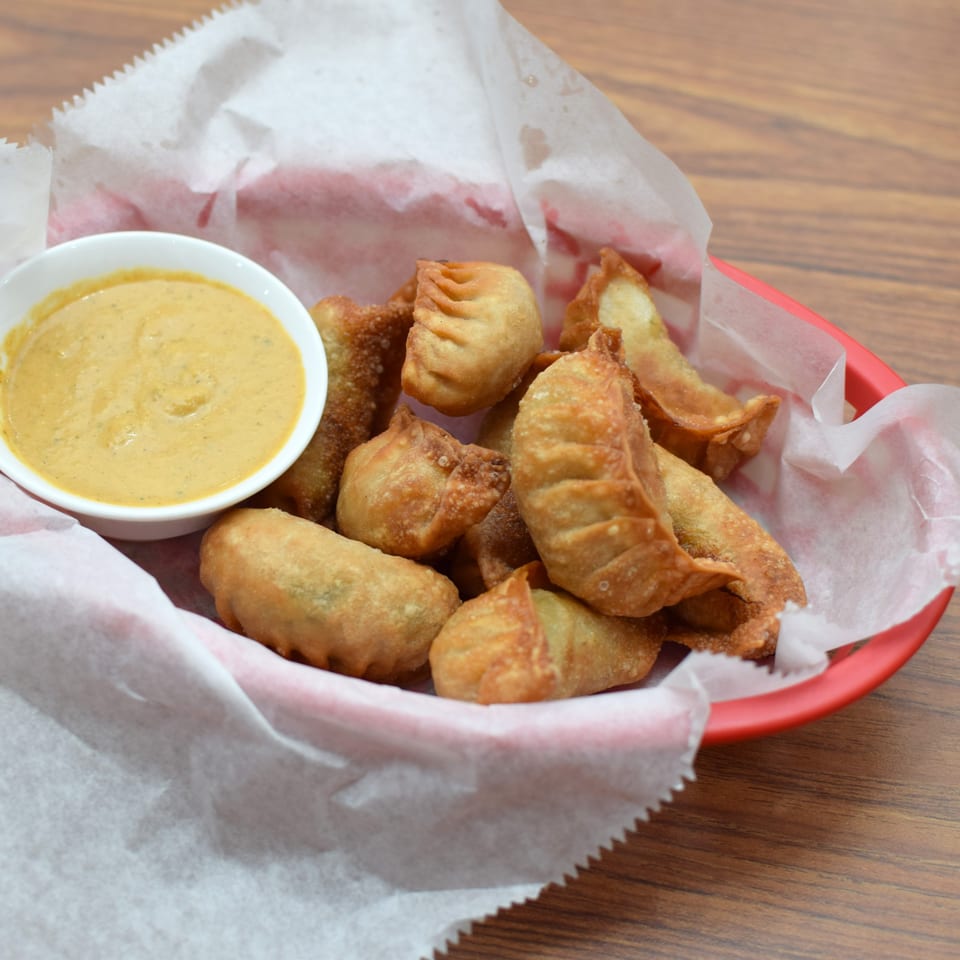 A red basket of crispy fried momos with a side of creamy dipping sauce on a wooden table.