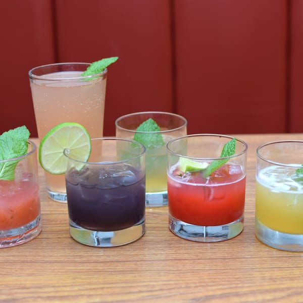 Six colorful Indian-inspired cocktails, garnished with mint and lime, on a wooden table.
