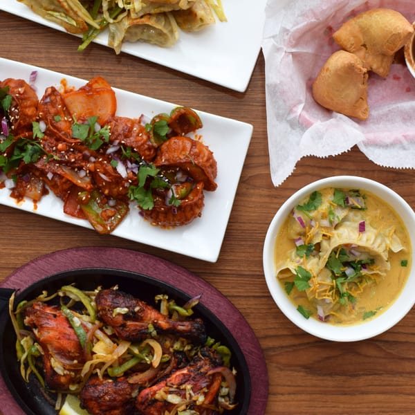 An overhead view of various Indian dishes, including chicken, momos, and samosas, on a wooden table.