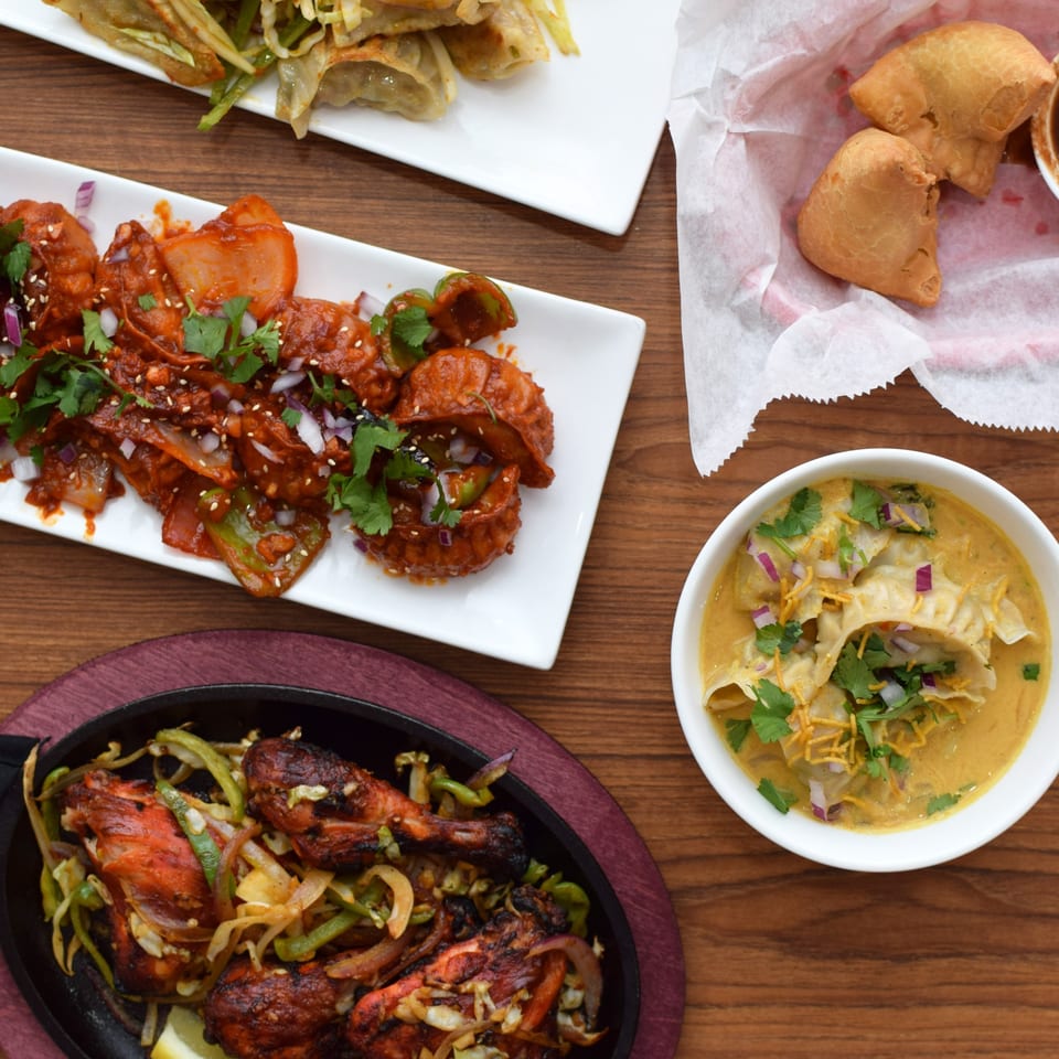 An overhead view of various Indian dishes, including chicken, momos, and samosas, on a wooden table.